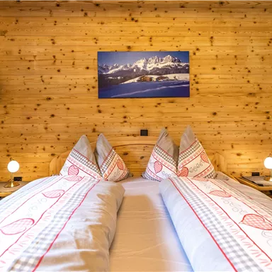 A cozy bedroom with two beds and wood paneling. A picture of a mountain landscape hangs on the wall and there are stylish bedside lamps.