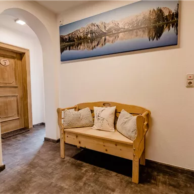 An inviting hallway with a wooden sofa and cushions. On the wall hangs a large picture of a mountain landscape mirrored calmly in the water.
