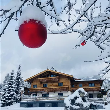 A snow-covered landscape image with a pretty wooden house. In the foreground, a red Christmas ornament hangs from a tree.