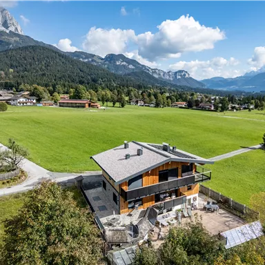 A modern house in a rural setting with a view of the mountains. The property is surrounded by a large, green meadow.