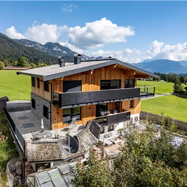 A modern wooden house in a picturesque landscape. In the background, there are mountains and a blue sky with clouds.