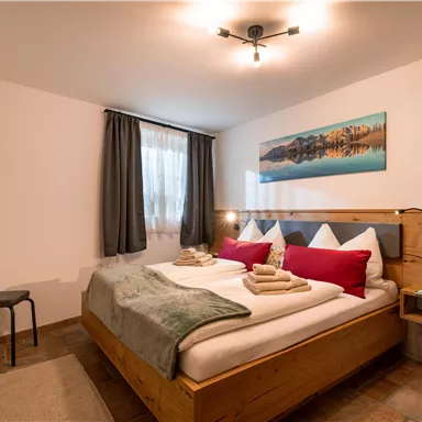 A modern bedroom with a double bed and wood-colored furniture. The room is bright with a large window and a television on the wall.