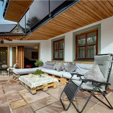 A cozy terrace with seating and a modern wooden overhang. The surroundings are bright and inviting, perfect for relaxation.