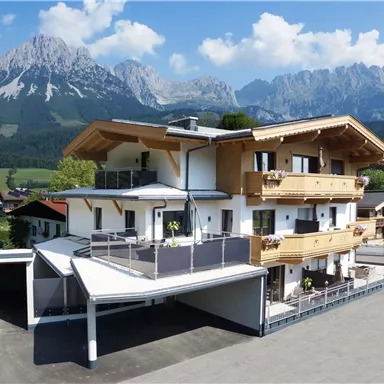 Ein modernes Chalet im Alpenstil mit großen Balkonen. Im Hintergrund sind majestätische Berge und eine grüne Landschaft zu sehen.