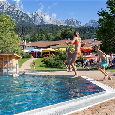 Ein Schwimmbad mit spritzenden Kindern und einer schönen Berglandschaft im Hintergrund. Die Sonne scheint, und die Umgebung ist lebhaft und einladend.