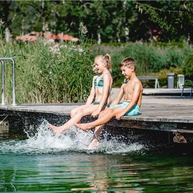 Zwei Kinder sitzen auf einem Steg am Wasser und lassen ihre Füße ins Wasser plätschern. Umgeben von Gras und Bäumen genießen sie den Tag.