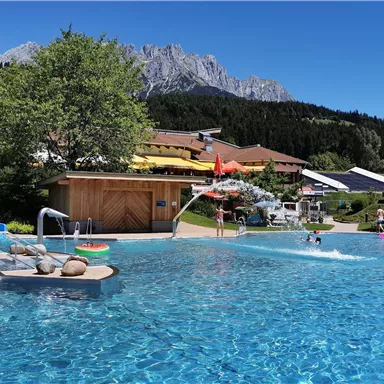A beautiful pool with clear water and a sunny surroundings. In the background, mountains and green areas can be seen.
