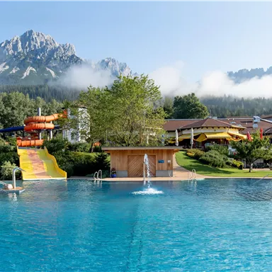 A beautiful pool with clear water and a giant slide. In the background, majestic mountains and green trees can be seen.