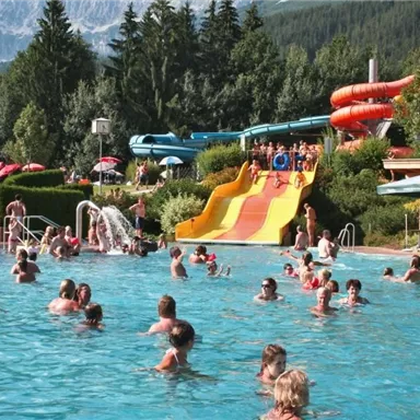 Ein belebter Schwimmbad mit vielen Menschen im Wasser. Im Hintergrund sieht man eine Rutsche und Grünanlagen.