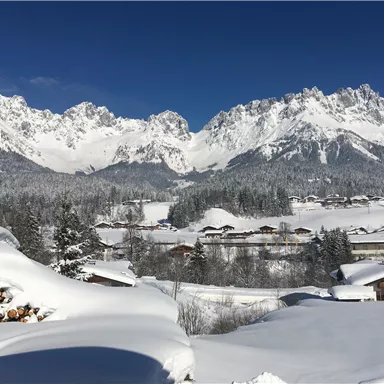 Eine winterliche Landschaft mit schneebedeckten Bergen und einem klaren blauen Himmel. Im Vordergrund sind verschneite Dächer und Holzscheite zu sehen.