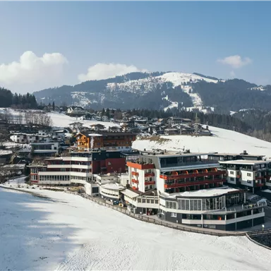 A picturesque mountain landscape with snow-covered hills. In the foreground, modern buildings are visible near a ski slope.