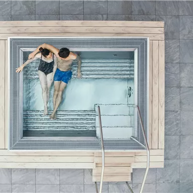 A couple sits relaxed in a modern hot tub. The area is brightly and invitingly designed, surrounded by gray tiles.