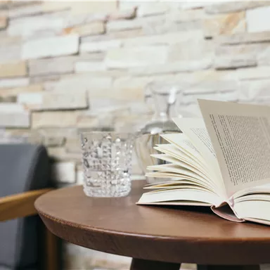 An open book lies on a round wooden table. Next to the book are a glass of water and a jug, in front of a beautiful stone wall.