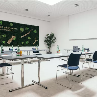 A modern meeting room with tables and chairs. On the wall hangs a green decorative panel with plant motifs and an inspiring quote.