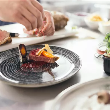 A filled plate with filleted meat presentation and colorful garnish. In the background, additional ingredients and utensils for preparation are visible.