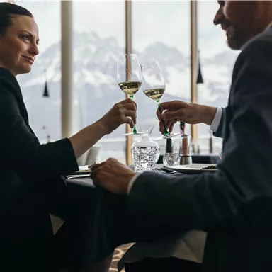 A couple toasts with champagne glasses in an elegant restaurant. Snow-covered mountains can be seen in the background.