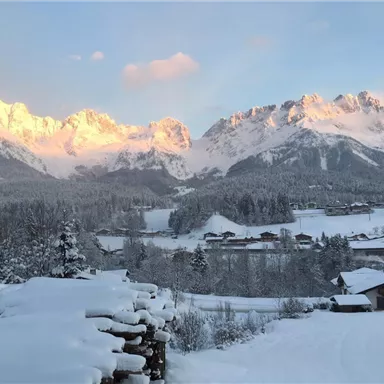 A wintry landscape with snow-covered mountains in the background. Small houses are visible nearby, while the sun illuminates the mountain peaks.
