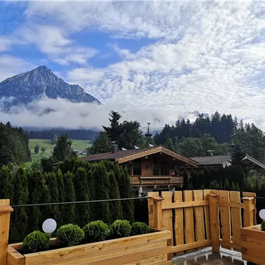 A picturesque mountain landscape with tall peaks and a cloudy sky. In the foreground, a wooden terrace with green plants and a view of adjacent houses.