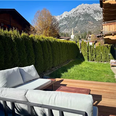 A quiet garden with a wooden terrace and comfortable furniture. In the background, green hedges and mountains can be seen.