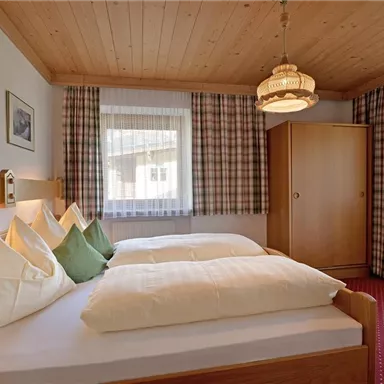A cozy bedroom with two beds and wooden furniture. The windows are fitted with curtains, and the walls are painted in warm colors.