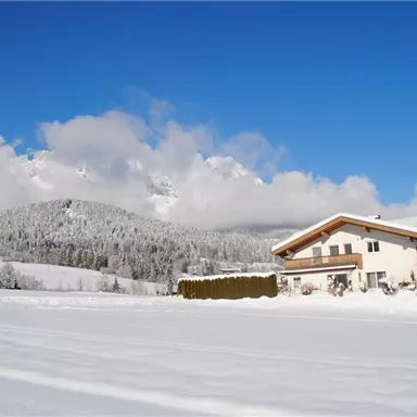 A snowy landscape with a charming house and majestic mountains in the background. The sky is clear and blue, creating a serene winter atmosphere.