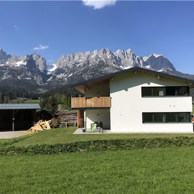 Ein modernes Haus in einer malerischen Berglandschaft. Im Hintergrund sind beeindruckende Berge und ein klarer blauer Himmel zu sehen.
