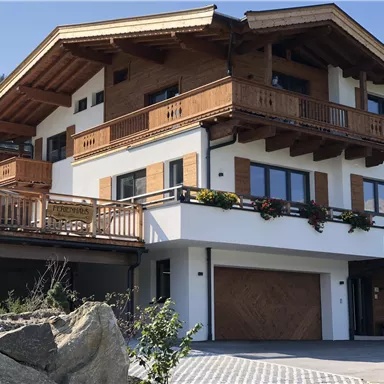 Ein modernes Haus im alpinen Stil mit Holzverkleidung und großen Balkonen. Die Umgebung ist gepflegt und es gibt eine Auffahrt mit Steinen.