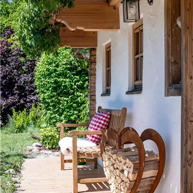 A cozy porch with a wooden chair and a wooden heart for firewood. In the background, green plants and a beautiful garden can be seen.