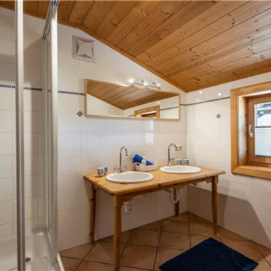 A modern bathroom with a shower and a double sink. Wooden ceilings and large windows create a bright, friendly atmosphere.