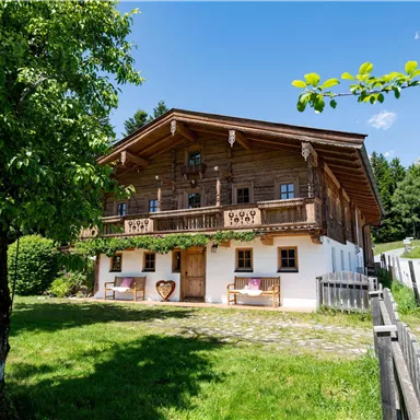 A traditional wooden house with a balcony in a green environment. It is a sunny day with a blue sky and trees in the foreground.