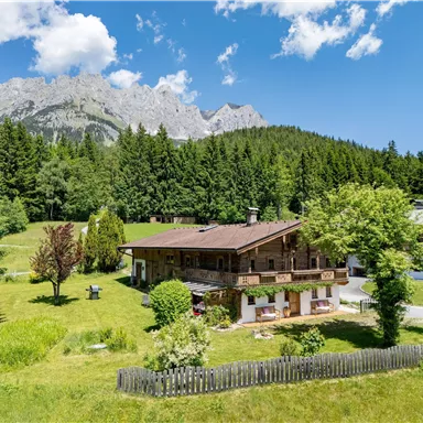 A picturesque alpine landscape with a wooden house and lush greenery. In the background, impressive mountains rise under a blue sky.