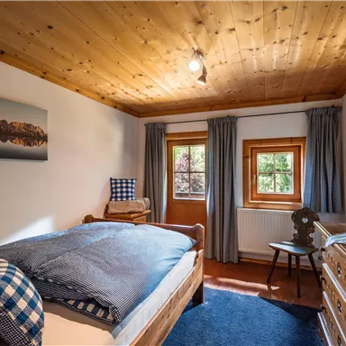 A cozy bedroom with a wooden ceiling and large windows. There is a single bed, a chair, and a dresser as well as a landscape photograph on the wall.