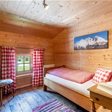 A cozy room with wooden walls and a single bed. Red checkered curtains and a beautiful view of the mountains complement the rustic atmosphere.