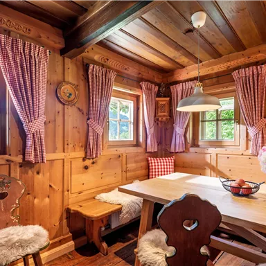 A cozy wooden cottage with a rustic dining table and chairs. The walls are made of wood, and the windows are adorned with checkered curtains.