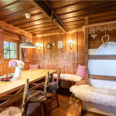 A cozy room with wooden walls and a large table. The furnishings are rustic, with windows, wooden lamps, and plaid-patterned curtains.
