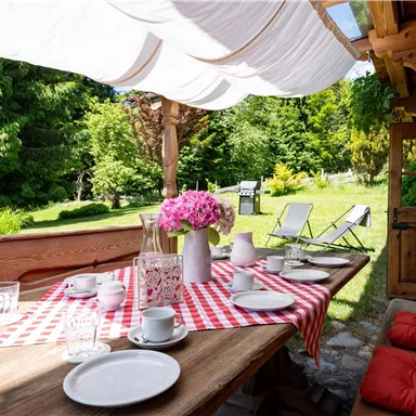 An inviting outdoor dining table with a red check pattern. The table is decorated with dishes and fresh flowers, surrounded by a green garden.