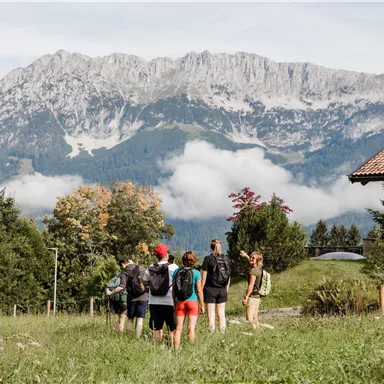 A group of hikers enjoys the view of mountains and a picturesque landscape. In the foreground, a beautiful, rustic house can be seen.