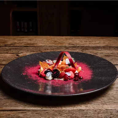 An artistically arranged dessert plate on a black plate. The decoration consists of various fruits, a crispy garnish, and a colorful sauce.