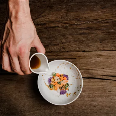 A hand pours sauce into a white bowl with colorful ingredients. The background consists of a wooden board.