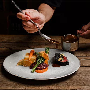 A plate with artfully arranged food, consisting of meat, vegetables, and various side dishes. A hand is pouring sauce over the dish.