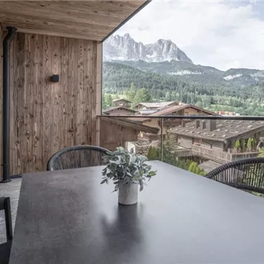 Ein moderner Balkon mit Holzwänden und einem Tisch. Im Hintergrund sind majestätische Berge und das grüne Tal sichtbar.