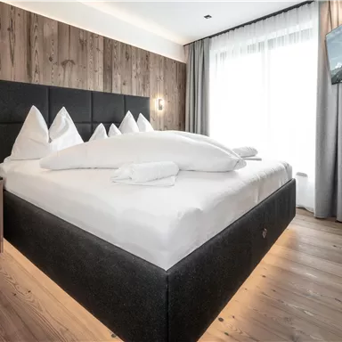 Ein modernes Schlafzimmer mit einem großen Bett und weißen Kissen. Die Wände sind mit Holz verkleidet, und es gibt einen Fernseher sowie große Fenster, die viel Licht hereinlassen.
