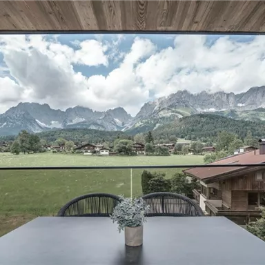 Eine beeindruckende Berglandschaft mit grünen Wiesen und Häusern im Vordergrund. Der Himmel ist bewölkt, und die Berge sind majestätisch im Hintergrund zu sehen.