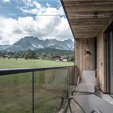 Ein Balkon mit Liegen und Blick auf hohe Berge. Im Hintergrund sind grüne Wiesen und ein bewölkter Himmel zu sehen.
