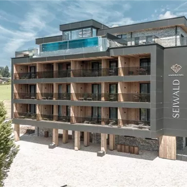 Ein modernes Hotelgebäude mit Holzelementen und großen Fenstern. Im Hintergrund sind Berge und eine Grünfläche zu sehen.