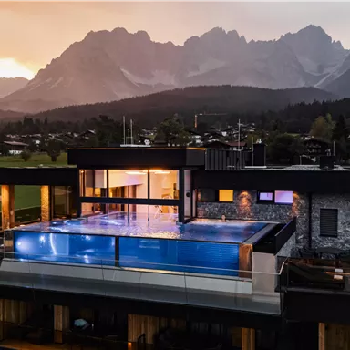 Ein modernes Haus mit einem Infinity-Pool und Blick auf die Berge. Die Szenerie wird von einem beeindruckenden Sonnenuntergang beleuchtet.