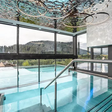 Ein modernes Schwimmbad mit Glasfront und Blick auf grüne Hügel. Das Wasser reflektiert die Umgebung und schafft eine entspannende Atmosphäre.