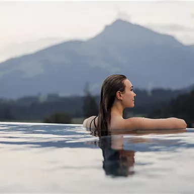 Eine Frau entspannt in einem Infinity-Pool mit Bergblick. Die sanfte Abendstimmung und die ruhige Landschaft schaffen eine entspannende Atmosphäre.