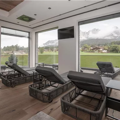 Ein moderner Raum mit Liegen und großen Fenstern, die eine grüne Landschaft und Berge zeigen. Der Raum wirkt hell und einladend.