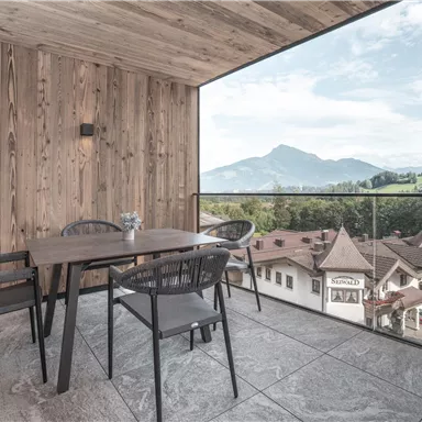 Ein modern eingerichteter Balkon mit einem Holzboden und einem Tisch aus Holz. Im Hintergrund sind Berge und eine idyllische Stadtlandschaft zu sehen.